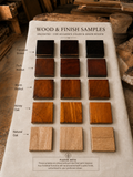 A white sample board labeled 'WOOD & FINISH SAMPLES' showing multiple wood finish swatches with handwritten color and grain reviews. The swatches include Espresso Brown, Dark Walnut, Warm Walnut, Honey Oak, and Natural Oak, arranged in rows on a workshop table with wood pieces in the background.